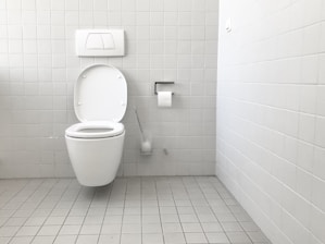 A clean and minimalistic bathroom featuring a wall-mounted toilet with an open lid. The walls and floor are tiled in a light gray color. A roll of toilet paper is mounted on the wall nearby, accompanied by a toilet brush in a holder.