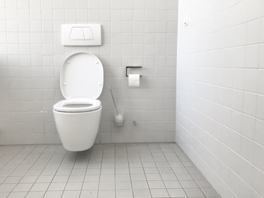 A clean and minimalistic bathroom featuring a wall-mounted toilet with an open lid. The walls and floor are tiled in a light gray color. A roll of toilet paper is mounted on the wall nearby, accompanied by a toilet brush in a holder.