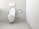 A clean and minimalistic bathroom featuring a wall-mounted toilet with an open lid. The walls and floor are tiled in a light gray color. A roll of toilet paper is mounted on the wall nearby, accompanied by a toilet brush in a holder.