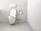 A clean and minimalistic bathroom featuring a wall-mounted toilet with an open lid. The walls and floor are tiled in a light gray color. A roll of toilet paper is mounted on the wall nearby, accompanied by a toilet brush in a holder.