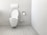 A clean and minimalistic bathroom featuring a wall-mounted toilet with an open lid. The walls and floor are tiled in a light gray color. A roll of toilet paper is mounted on the wall nearby, accompanied by a toilet brush in a holder.