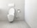 A clean and minimalistic bathroom featuring a wall-mounted toilet with an open lid. The walls and floor are tiled in a light gray color. A roll of toilet paper is mounted on the wall nearby, accompanied by a toilet brush in a holder.