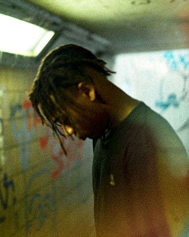 A person with dreadlocks is standing in an urban setting with graffiti on the walls. The lighting is dim and the atmosphere seems introspective and solitary.