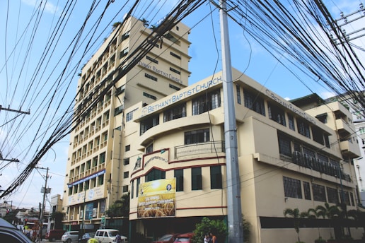 A large, multi-story building with clear signage indicating it is a Baptist church. Numerous power lines run across the foreground, intersecting in complex patterns. The sky is partly cloudy, and a few vehicles and people are visible on the street below.