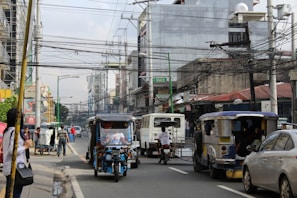 A busy urban street with several vehicles, including a jeepney and tricycles, driving through. People are walking on the sidewalk while others are sitting in the jeepney. Overhead, there is a dense network of electrical wires stretching across the road. The street is lined with buildings, and signs are visible in the background.