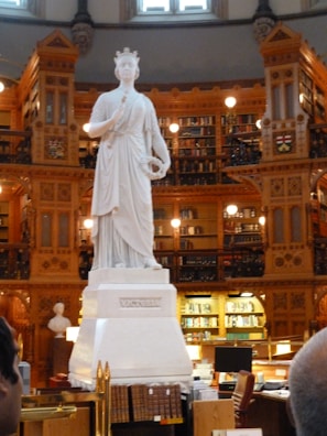A statue of a robed figure stands prominently in the center of an ornate library. The library features intricate woodwork, abundant bookshelves, and warm lighting. The base of the statue is inscribed with 'Victoria'.