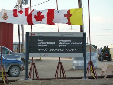 A sign for the Polar Continental Shelf Program by Natural Resources Canada is prominently displayed. The sign includes English and Inuktitut languages. Above the sign, several flags wave in the wind, including the Canadian flag. The area appears to be an outdoor setting with construction materials and vehicles visible.