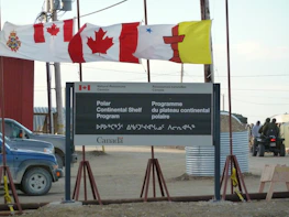 A sign for the Polar Continental Shelf Program by Natural Resources Canada is prominently displayed. The sign includes English and Inuktitut languages. Above the sign, several flags wave in the wind, including the Canadian flag. The area appears to be an outdoor setting with construction materials and vehicles visible.