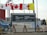 A sign for the Polar Continental Shelf Program by Natural Resources Canada is prominently displayed. The sign includes English and Inuktitut languages. Above the sign, several flags wave in the wind, including the Canadian flag. The area appears to be an outdoor setting with construction materials and vehicles visible.