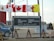 A sign for the Polar Continental Shelf Program by Natural Resources Canada is prominently displayed. The sign includes English and Inuktitut languages. Above the sign, several flags wave in the wind, including the Canadian flag. The area appears to be an outdoor setting with construction materials and vehicles visible.
