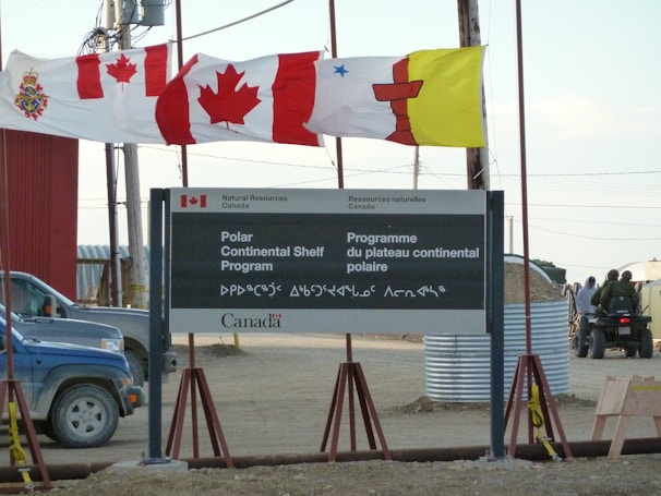 A sign for the Polar Continental Shelf Program by Natural Resources Canada is prominently displayed. The sign includes English and Inuktitut languages. Above the sign, several flags wave in the wind, including the Canadian flag. The area appears to be an outdoor setting with construction materials and vehicles visible.