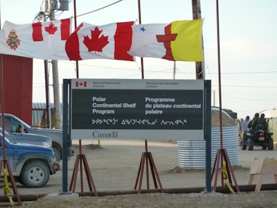 A sign for the Polar Continental Shelf Program by Natural Resources Canada is prominently displayed. The sign includes English and Inuktitut languages. Above the sign, several flags wave in the wind, including the Canadian flag. The area appears to be an outdoor setting with construction materials and vehicles visible.
