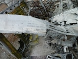A close-up view of the twisted and damaged remains of an aircraft, displaying torn metal structures and scattered debris. The structure shows visible text that has been partially obscured and surrounding metallic surfaces appear weathered and rusty.