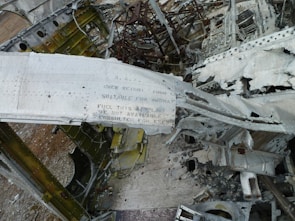 A close-up view of the twisted and damaged remains of an aircraft, displaying torn metal structures and scattered debris. The structure shows visible text that has been partially obscured and surrounding metallic surfaces appear weathered and rusty.