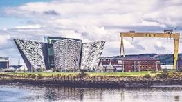 An architectural structure with a modern, geometric design featuring angular, metallic panels. Nearby, an industrial crane labeled 'H & W' stands prominently, and a red-brick building is visible in the background. The scene is reflected in the calm waters of the foreground, under a partly cloudy sky.