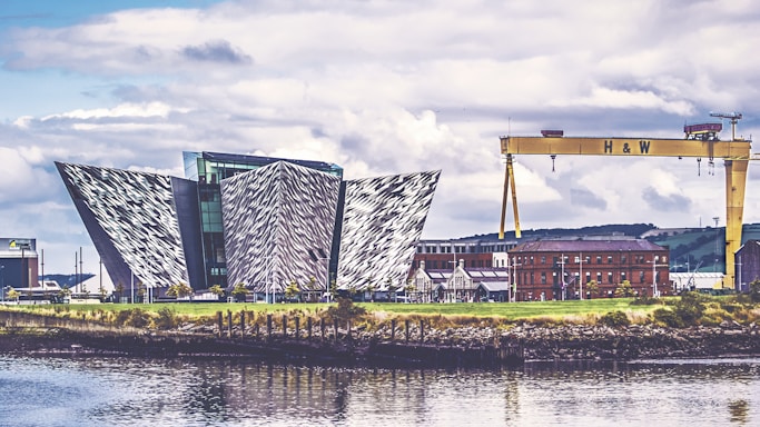 An architectural structure with a modern, geometric design featuring angular, metallic panels. Nearby, an industrial crane labeled 'H & W' stands prominently, and a red-brick building is visible in the background. The scene is reflected in the calm waters of the foreground, under a partly cloudy sky.