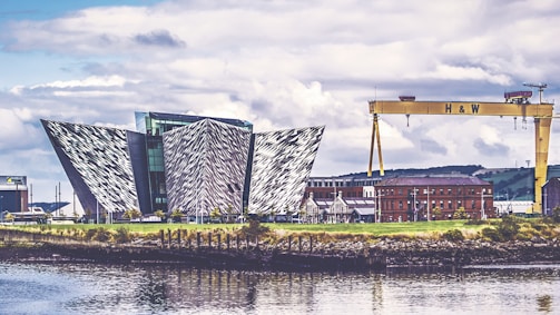 An architectural structure with a modern, geometric design featuring angular, metallic panels. Nearby, an industrial crane labeled 'H & W' stands prominently, and a red-brick building is visible in the background. The scene is reflected in the calm waters of the foreground, under a partly cloudy sky.