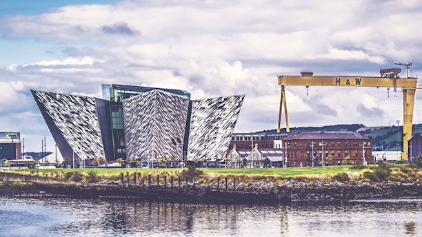 An architectural structure with a modern, geometric design featuring angular, metallic panels. Nearby, an industrial crane labeled 'H & W' stands prominently, and a red-brick building is visible in the background. The scene is reflected in the calm waters of the foreground, under a partly cloudy sky.