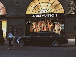 A luxury car is parked in front of a brightly lit Louis Vuitton store with an elaborate window display featuring colorful decorations. Two individuals walk on the street in front of the store. The storefront is framed by a large arched window, and there are additional shops seen on either side.