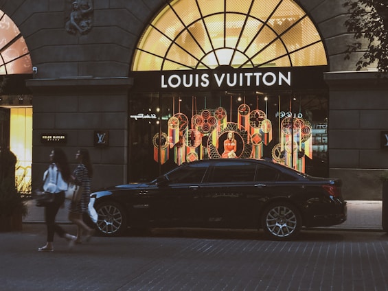 A luxury car is parked in front of a brightly lit Louis Vuitton store with an elaborate window display featuring colorful decorations. Two individuals walk on the street in front of the store. The storefront is framed by a large arched window, and there are additional shops seen on either side.