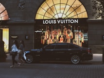 A luxury car is parked in front of a brightly lit Louis Vuitton store with an elaborate window display featuring colorful decorations. Two individuals walk on the street in front of the store. The storefront is framed by a large arched window, and there are additional shops seen on either side.