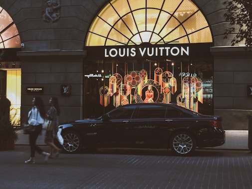 A luxury car is parked in front of a brightly lit Louis Vuitton store with an elaborate window display featuring colorful decorations. Two individuals walk on the street in front of the store. The storefront is framed by a large arched window, and there are additional shops seen on either side.