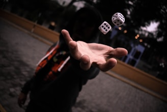 A person wearing a dark hoodie extends their hand forward as two dice are captured mid-air, creating a sense of motion and suspense. The background is slightly blurred, highlighting the focused hand and floating dice.