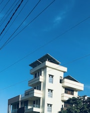 A modern multi-story building with rectangular windows and balconies, featuring solar panels on the rooftop. The sky is clear and blue, intersected by several power lines. Trees partially frame the lower part of the image.