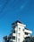 A modern multi-story building with rectangular windows and balconies, featuring solar panels on the rooftop. The sky is clear and blue, intersected by several power lines. Trees partially frame the lower part of the image.