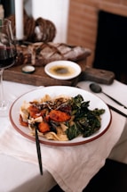 A plate of pasta served with roasted sweet potatoes, leafy greens, and a sprinkling of seeds on a white tablecloth. A glass of red wine stands nearby, and in the background, there is a loaf of rustic bread, a dish with olive oil, and a candle. The setting appears homely and inviting, with warm lighting.