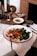 A plate of pasta served with roasted sweet potatoes, leafy greens, and a sprinkling of seeds on a white tablecloth. A glass of red wine stands nearby, and in the background, there is a loaf of rustic bread, a dish with olive oil, and a candle. The setting appears homely and inviting, with warm lighting.