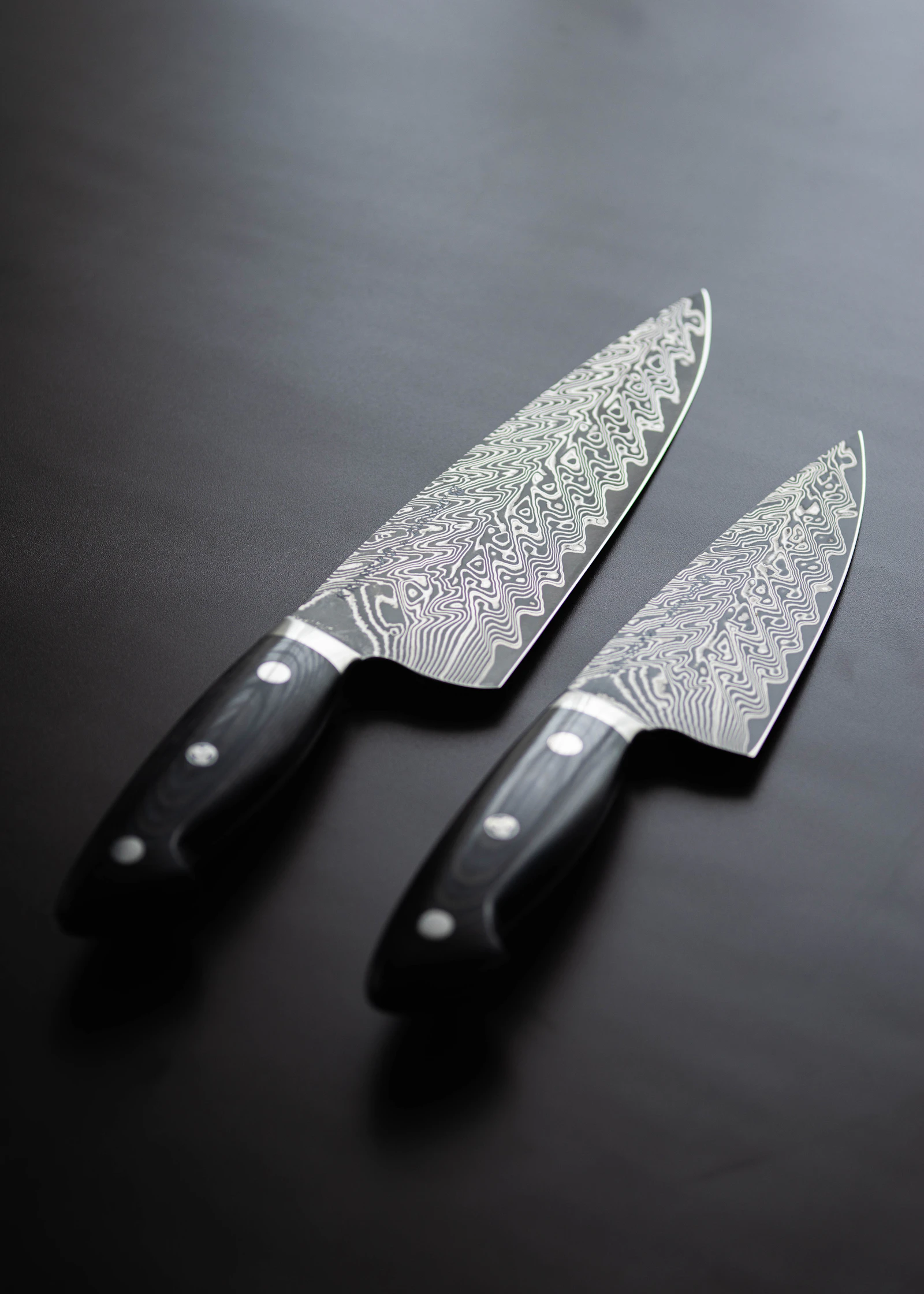 Two Damascus chef's knives with black riveted handles on a dark surface