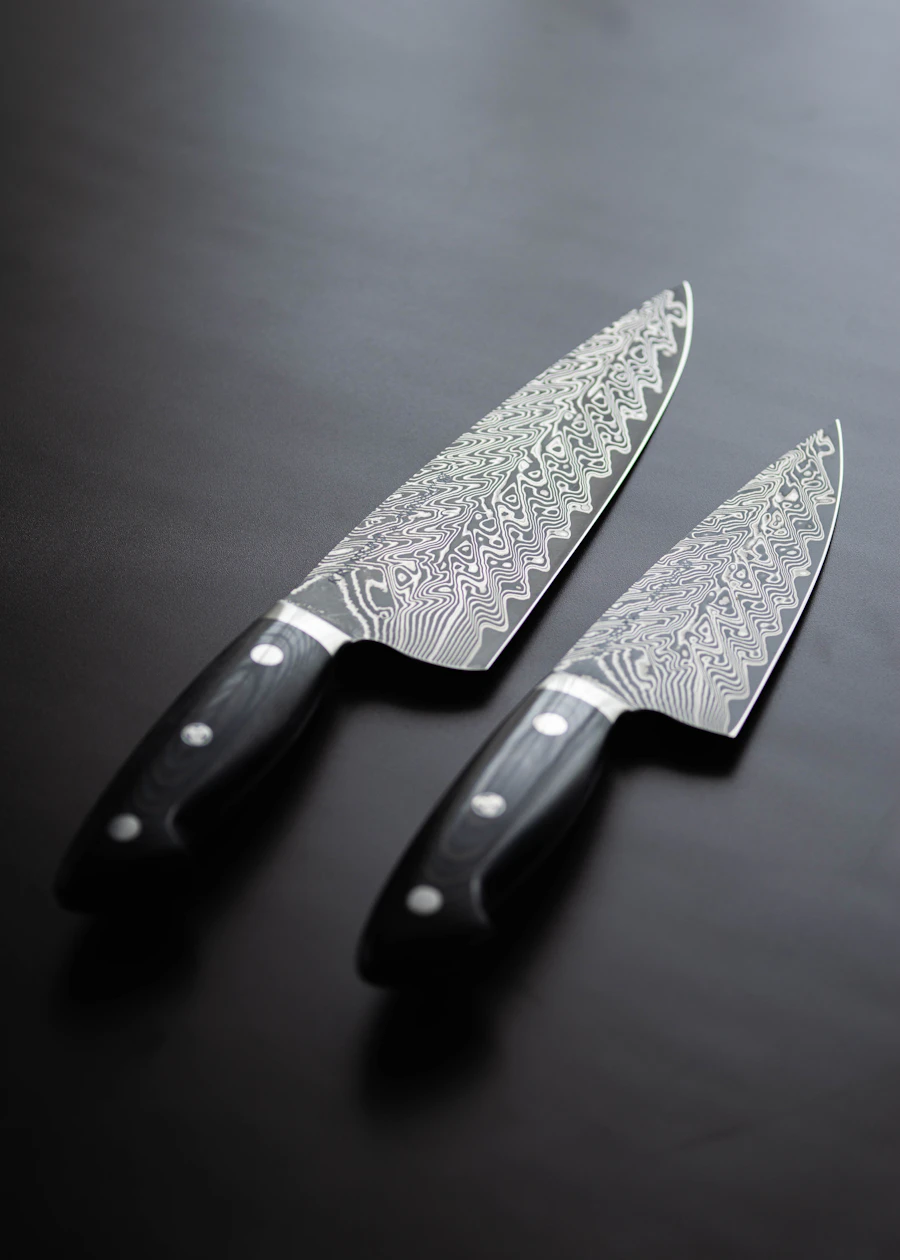 Two Damascus chef's knives with black riveted handles on a dark surface