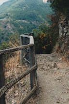 A narrow, rocky path with a wooden railing lined with wire mesh, set against a backdrop of dense green hills. The trail appears rugged and natural, with dry grass and scattered stones along the path. Lush foliage in varying shades of green and a hint of brown surrounds the area.