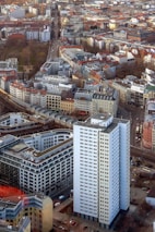 An aerial view of a densely packed urban area featuring a mix of modern and historic architecture. A prominent white high-rise building stands in the foreground, surrounded by a variety of smaller buildings with red and gray roofs. Streets are interwoven between the structures, with visible vehicles and greenery interspersed throughout the cityscape.