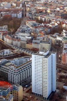 An aerial view of a densely packed urban area featuring a mix of modern and historic architecture. A prominent white high-rise building stands in the foreground, surrounded by a variety of smaller buildings with red and gray roofs. Streets are interwoven between the structures, with visible vehicles and greenery interspersed throughout the cityscape.