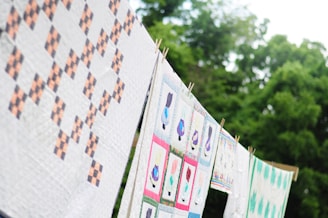 Brightly colored quilts hang on a clothesline against a backdrop of lush, green trees. The designs feature geometric patterns and playful images, showcasing a variety of shapes and hues.