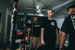 Three men are gathered in a room with film equipment, appearing to work together. They are casually dressed, one wearing a shirt with '4K' printed on it. The environment is dimly lit with a ceiling fan above and some board games like Trivial Pursuit visible in the background.