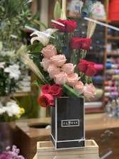 A tall arrangement of red and pink roses is complemented by white lilies and decorative peacock feathers. The flowers are arranged in a sleek black vase labeled 'Flower' and are displayed on a wooden stand in a floral shop setting.