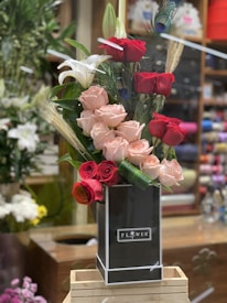 A tall arrangement of red and pink roses is complemented by white lilies and decorative peacock feathers. The flowers are arranged in a sleek black vase labeled 'Flower' and are displayed on a wooden stand in a floral shop setting.