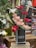 A tall arrangement of red and pink roses is complemented by white lilies and decorative peacock feathers. The flowers are arranged in a sleek black vase labeled 'Flower' and are displayed on a wooden stand in a floral shop setting.
