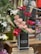 A tall arrangement of red and pink roses is complemented by white lilies and decorative peacock feathers. The flowers are arranged in a sleek black vase labeled 'Flower' and are displayed on a wooden stand in a floral shop setting.