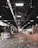 A large industrial warehouse with a high ceiling and ceiling fans. Stacks of compacted cardboard and paper are scattered throughout the space. Overhead lighting provides illumination, creating a contrast with the shadows. The floor is littered with loose debris.