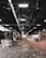 A large industrial warehouse with a high ceiling and ceiling fans. Stacks of compacted cardboard and paper are scattered throughout the space. Overhead lighting provides illumination, creating a contrast with the shadows. The floor is littered with loose debris.