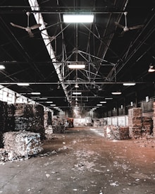 A large industrial warehouse with a high ceiling and ceiling fans. Stacks of compacted cardboard and paper are scattered throughout the space. Overhead lighting provides illumination, creating a contrast with the shadows. The floor is littered with loose debris.