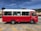 A red and white public transport minibus is parked in a lot on a sunny day. The vehicle has large windows with curtains and the text 'Public Transport Vehicle' is visible on the side. The parking area has yellow lines, and there's a background of trees and buildings under a clear blue sky.