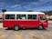 A red and white public transport minibus is parked in a lot on a sunny day. The vehicle has large windows with curtains and the text 'Public Transport Vehicle' is visible on the side. The parking area has yellow lines, and there's a background of trees and buildings under a clear blue sky.