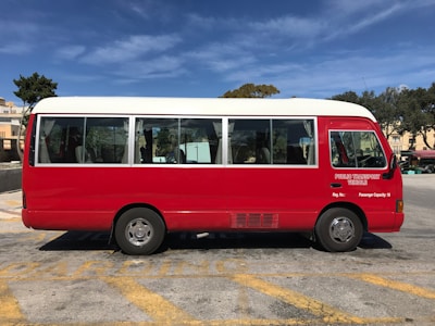 A red and white public transport minibus is parked in a lot on a sunny day. The vehicle has large windows with curtains and the text 'Public Transport Vehicle' is visible on the side. The parking area has yellow lines, and there's a background of trees and buildings under a clear blue sky.