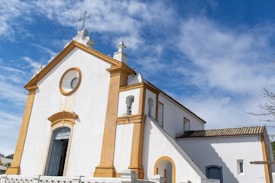A white church with golden yellow accents under a bright blue sky with wispy clouds. The building has a cross on top and arched doors and windows, and a staircase leading to an entrance.