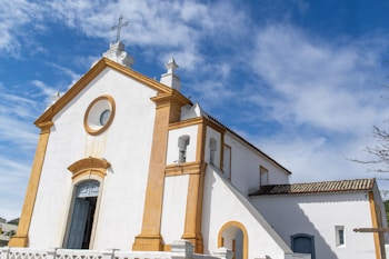 A white church with golden yellow accents under a bright blue sky with wispy clouds. The building has a cross on top and arched doors and windows, and a staircase leading to an entrance.